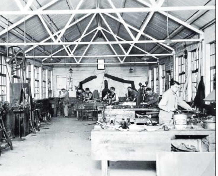 Black and white archive photo of a workshop with high steel-trussed ceilings, showing several workers at benches and machinery, connected to Emily Kathleen Renshaw's history.